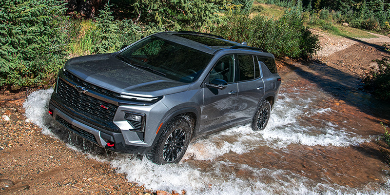 A dark gray Chevrolet Equinox SUV drives through a shallow stream on a dirt road, with trees in the background.