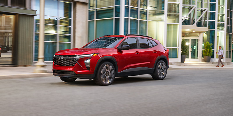 Red 2024 Chevrolet Trax RS driving on a city street past modern buildings, with a person walking on the sidewalk.