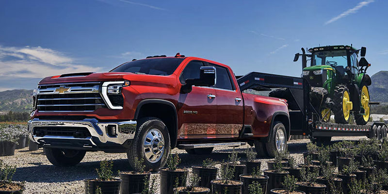 A red Chevrolet Silverado 3500 HD truck shown from the side, towing a green John Deere tractor on a trailer.