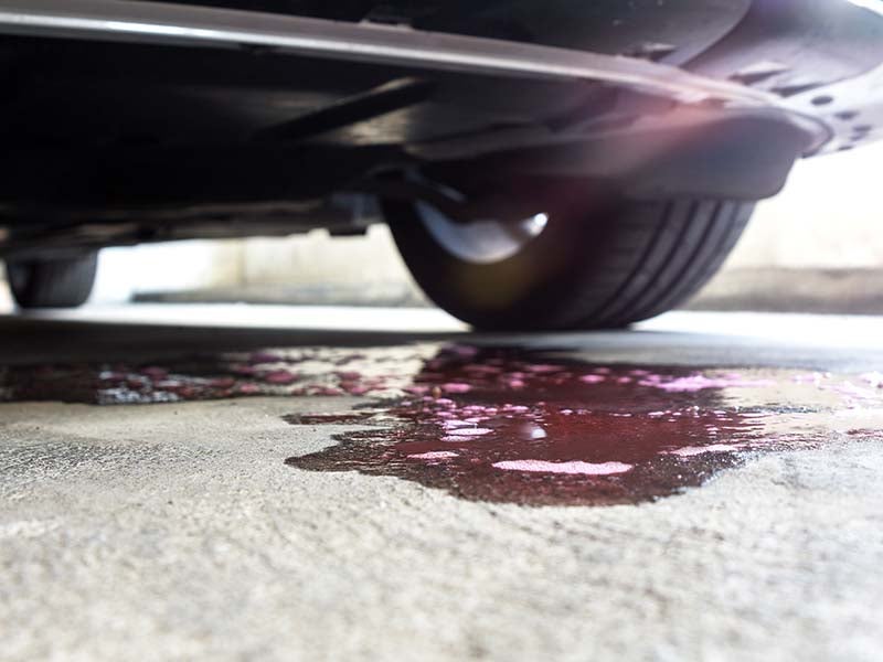 Pink-red liquid with bubbles leaking from under a car onto a concrete surface.