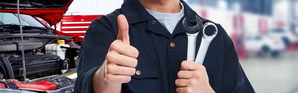 Auto mechanic in a blue uniform holds two wrenches and gives a thumbs up, with a car engine in the background.