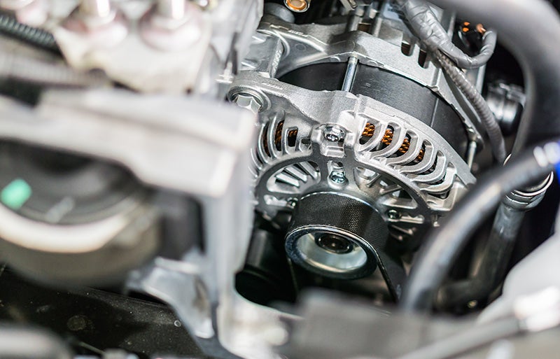 Close-up of a new car alternator with its pulley and drive belt, wires, and engine components.