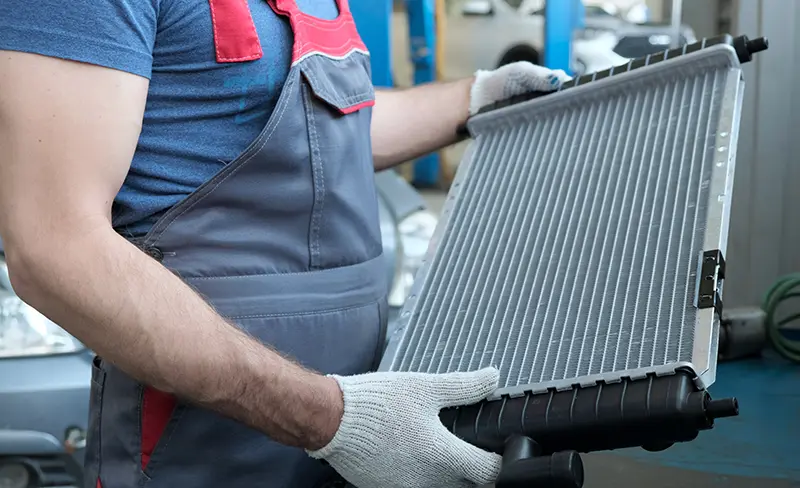 Mechanic in overalls and gloves holding up a new, silver car radiator in an auto repair shop.