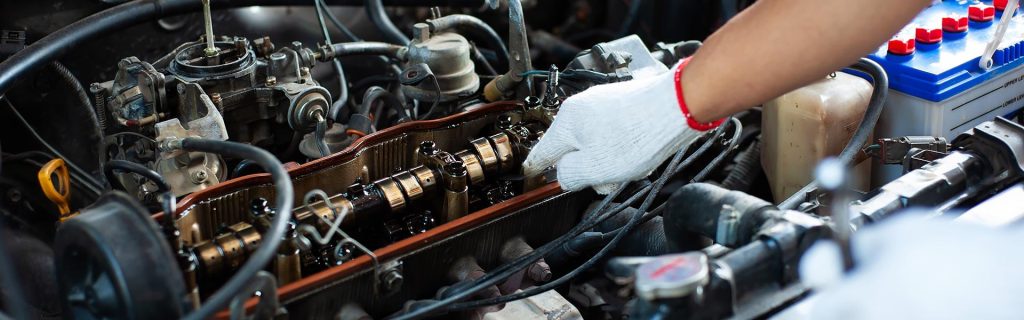 Gloved hand working on an exposed car engine with the valve cover removed, camshafts and rocker arms visible.