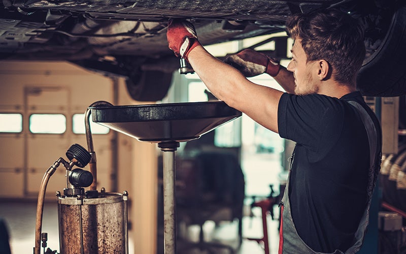 A mechanic in red gloves and a gray apron drains oil from a car high overhead into a large metal funnel.