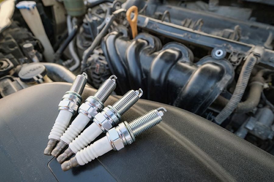 Four new spark plugs resting on a car's engine cover, with the engine itself blurred in the background.