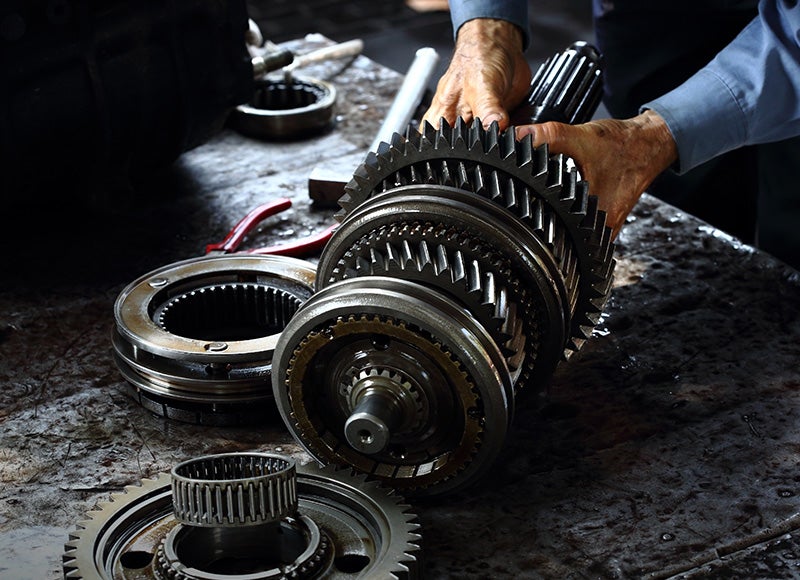 A mechanic's hands assemble a transmission gear set on a workbench with other disassembled parts.