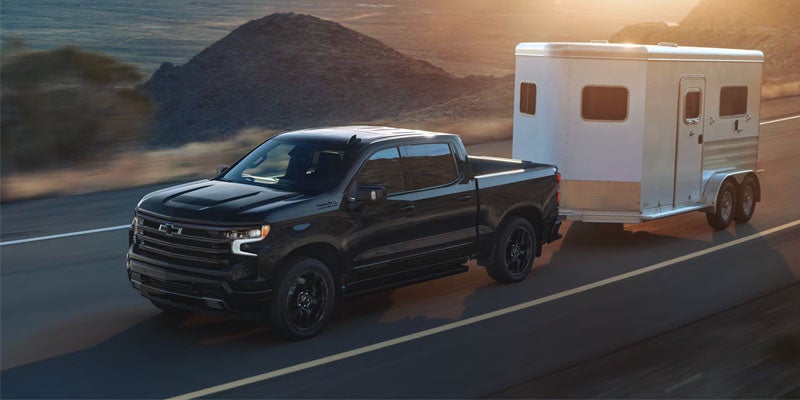 A black Chevrolet pickup truck tows a white travel trailer along a mountain road at sunset.