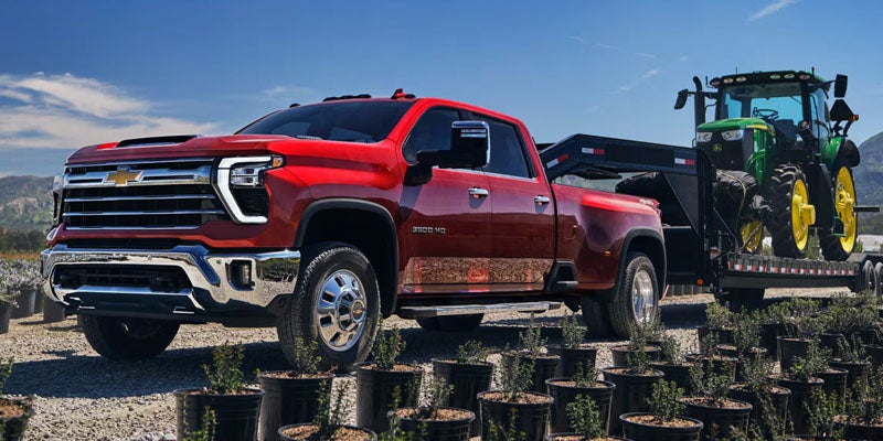 Chevrolet Silverado HD pickup truck towing heavy equipment with a tractor trailer outdoors.